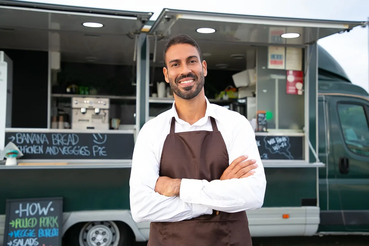 A food truck owner in a white shirt and brown apron stands smiling with his arms crossed in front of his dark green truck, serving window open. Chalkboards around him read BANANA BREAD, CHICKEN AND VEGGIES PIE, WE HAVE WI-FI, and a menu listing PULLED PORK, VEGGIE BURGER and SALADS.