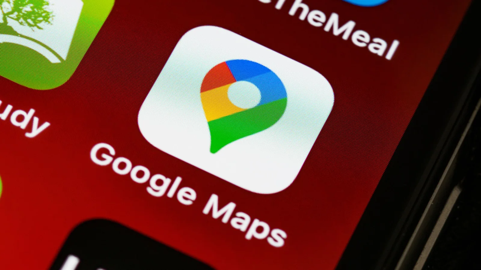 Close-up of a phone screen showing the Google Maps app icon, the coloured location pin on a white tile, labelled Google Maps, with ShareTheMeal above it and Uber below on a dark red background.